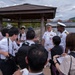 Two U.S. Navy officers in white dress uniforms are being interviewed or briefed by a group of Japanese civilians and journalists near a waterfront location, likely in Japan. The setting appears to be an outdoor area near a harbor or bay, with a wooden pavilion structure visible in the background. The interaction suggests a press engagement or community relations event.