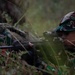 A soldier in camouflage uniform is lying prone in tall grass, aiming a rifle equipped with an optical scope. A second soldier is partially visible in the background. The setting appears to be a field training exercise based on the environment and equipment visible.
