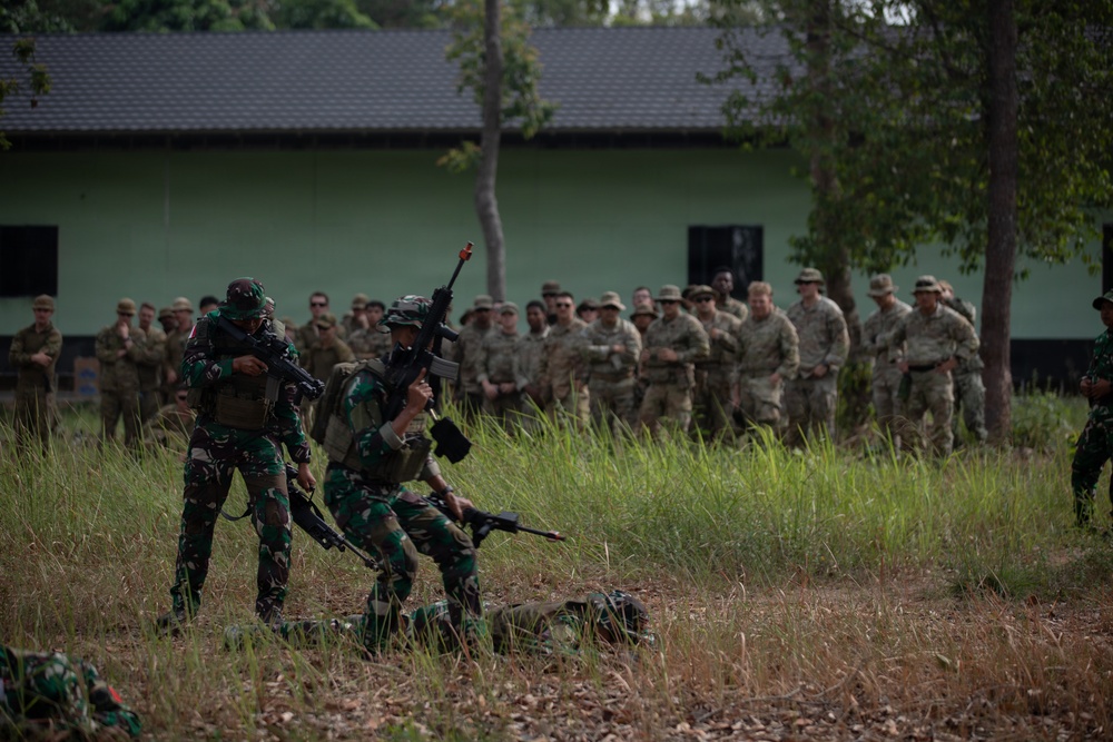 A military training exercise is taking place outdoors near a green building. Two soldiers in green camouflage uniforms (likely Indonesian TNI) are demonstrating a tactical drill — one raising a rifle overhead — while a large group of soldiers in tan/multicam uniforms observes from the background. The scene appears to be a joint military training event between two different national forces.