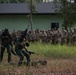 A military training exercise is taking place outdoors near a green building. Two soldiers in green camouflage uniforms (likely Indonesian TNI) are demonstrating a tactical drill — one raising a rifle overhead — while a large group of soldiers in tan/multicam uniforms observes from the background. The scene appears to be a joint military training event between two different national forces.