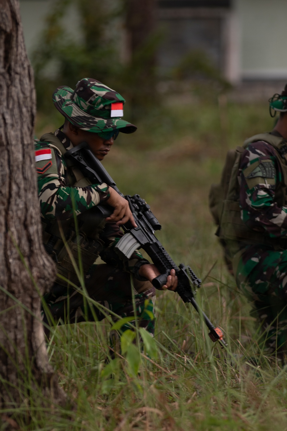 Two soldiers in Indonesian military (TNI - Tentara Nasional Indonesia) camouflage uniforms are positioned near a tree in a grassy field. The primary soldier in the foreground is crouching and holding an assault rifle, wearing a boonie hat with an Indonesian flag patch. The scene appears to depict a military training exercise or tactical field operation.