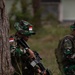 Two soldiers in Indonesian military (TNI - Tentara Nasional Indonesia) camouflage uniforms are positioned near a tree in a grassy field. The primary soldier in the foreground is crouching and holding an assault rifle, wearing a boonie hat with an Indonesian flag patch. The scene appears to depict a military training exercise or tactical field operation.