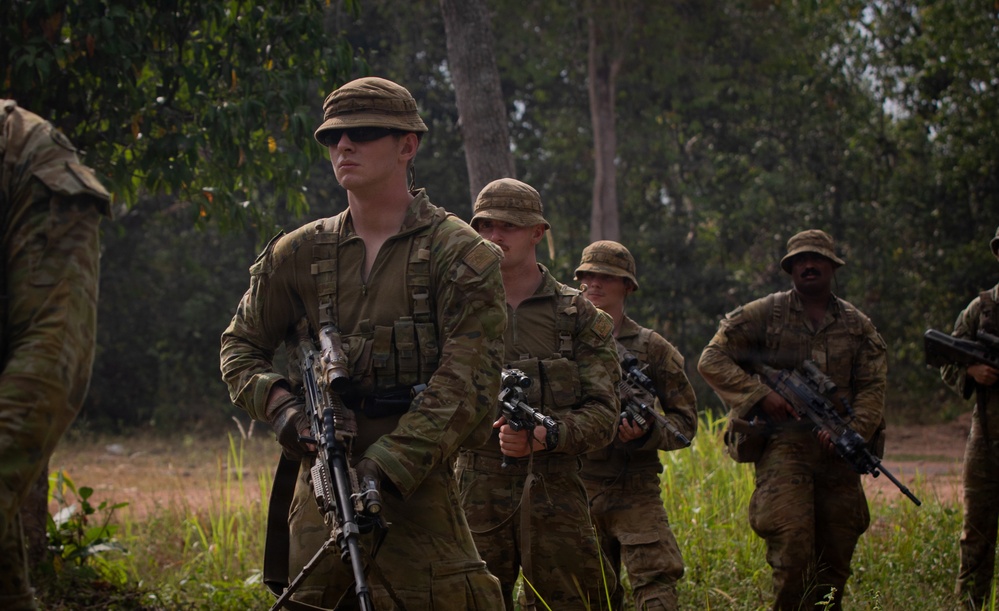 A group of soldiers in camouflage uniforms are conducting a patrol or field exercise in a tropical or subtropical outdoor environment. They are armed with assault rifles and wearing tactical gear including chest rigs and boonie hats. The lead soldier is wearing sunglasses and carrying his weapon at the low ready position.