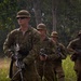 A group of soldiers in camouflage uniforms are conducting a patrol or field exercise in a tropical or subtropical outdoor environment. They are armed with assault rifles and wearing tactical gear including chest rigs and boonie hats. The lead soldier is wearing sunglasses and carrying his weapon at the low ready position.