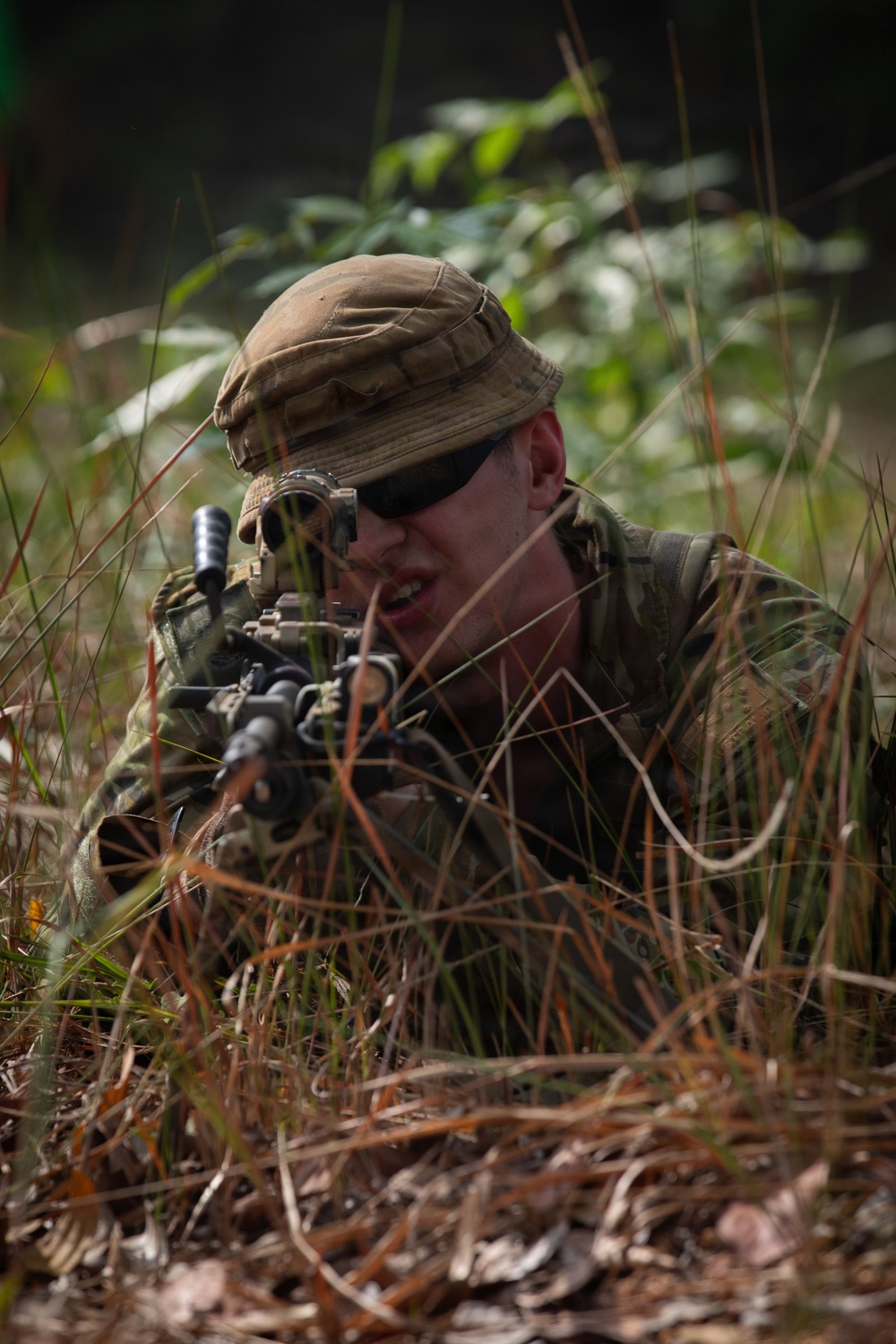 A soldier in full camouflage gear is lying prone in tall grass and vegetation, aiming a rifle forward in a low-profile tactical position. The individual is wearing a boonie hat, tactical sunglasses, and multicam/camouflage uniform, blending into the natural surroundings. The setting appears to be a wooded or jungle-edge environment, consistent with a military training exercise or field operation.