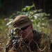 A soldier in full camouflage gear is lying prone in tall grass and vegetation, aiming a rifle forward in a low-profile tactical position. The individual is wearing a boonie hat, tactical sunglasses, and multicam/camouflage uniform, blending into the natural surroundings. The setting appears to be a wooded or jungle-edge environment, consistent with a military training exercise or field operation.