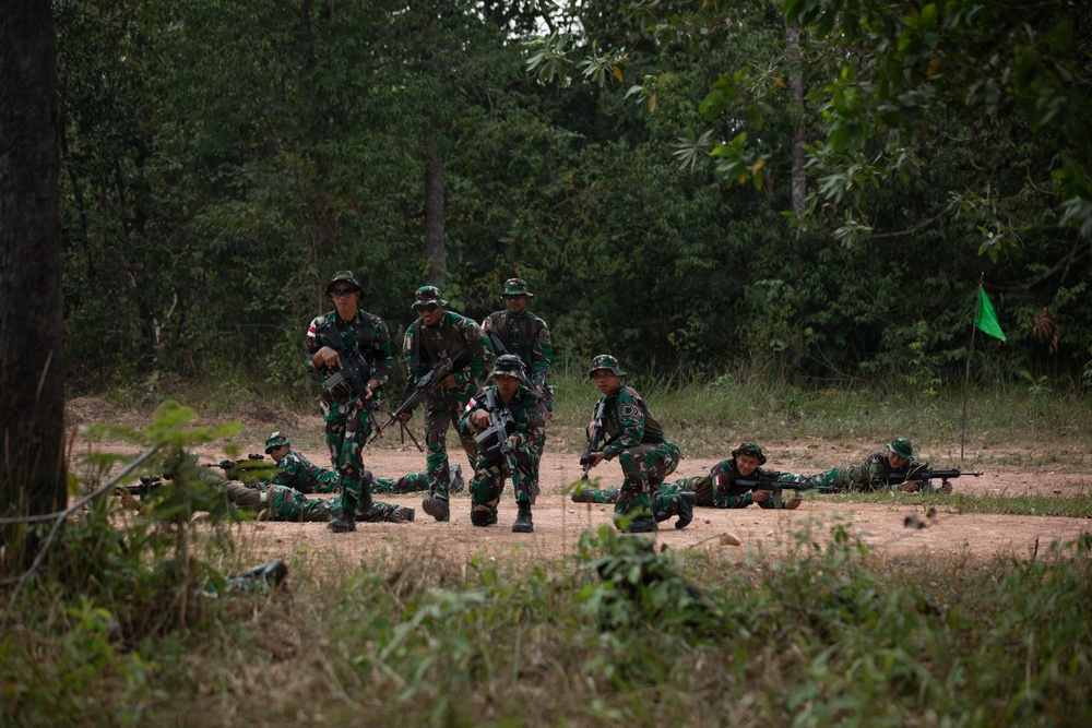 A group of soldiers in camouflage uniforms are conducting a military field training exercise in a forested clearing. Some personnel are in prone firing positions on the ground while others stand or crouch in tactical stances, all carrying rifles. A small green flag is visible on the right side, likely marking a position or boundary.
