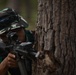 An Indonesian military soldier is positioned behind a large tree trunk, aiming a rifle with an optical sight attachment. The soldier is wearing woodland camouflage uniform and a boonie hat, with a visible 'Indonesia' patch and Indonesian flag on the left arm. This appears to be a military training exercise or tactical drill in a forested environment.