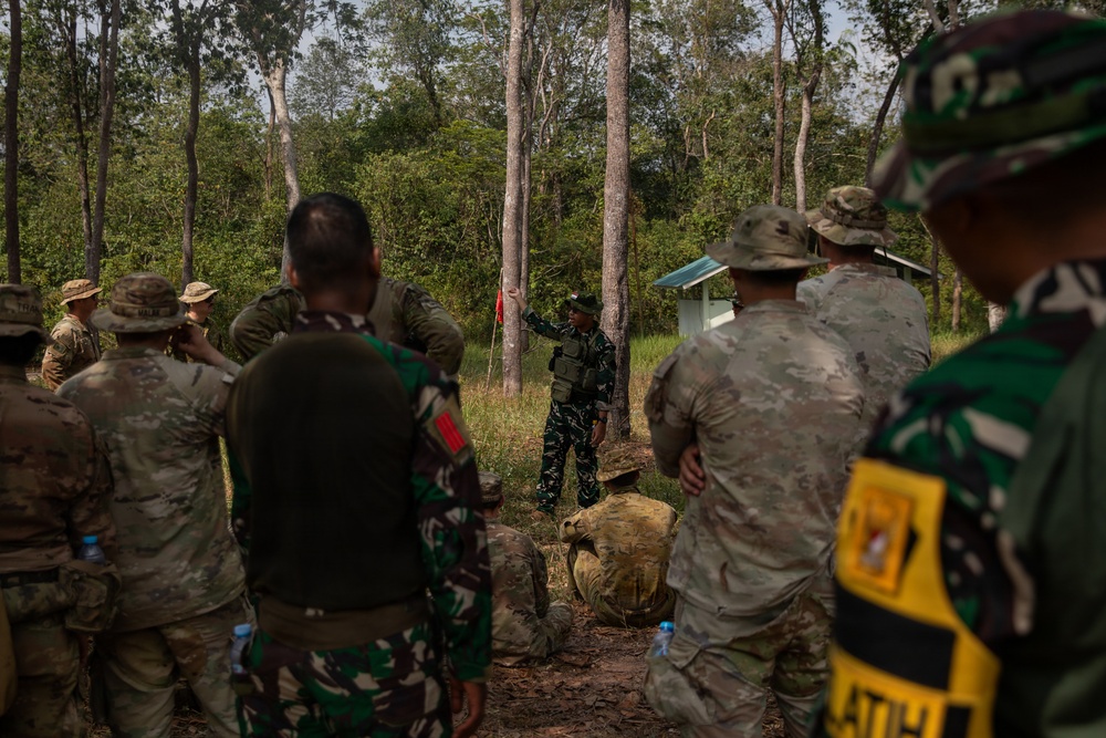 A joint military training exercise is taking place in a forested outdoor environment. Personnel in two distinct uniforms — US-style multicam/OCP camouflage and Indonesian TNI (Tentara Nasional Indonesia) camouflage — are gathered around an instructor who appears to be conducting a briefing or demonstration. A yellow patch reading 'LATIH' (Indonesian for 'training') is visible on one participant's vest, confirming this is a training exercise.