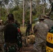 A joint military training exercise is taking place in a forested outdoor environment. Personnel in two distinct uniforms — US-style multicam/OCP camouflage and Indonesian TNI (Tentara Nasional Indonesia) camouflage — are gathered around an instructor who appears to be conducting a briefing or demonstration. A yellow patch reading 'LATIH' (Indonesian for 'training') is visible on one participant's vest, confirming this is a training exercise.