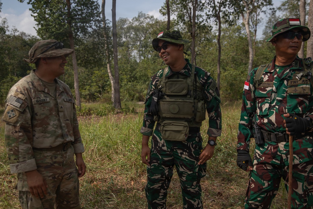 Three military personnel are standing together in an outdoor wooded area, appearing to be engaged in friendly conversation. One soldier is wearing US Army multicam uniform with a name tape reading 'VASQUEZ' and an American flag patch, while the other two are wearing Indonesian Army (TNI - Tentara Nasional Indonesia) camouflage uniforms with Indonesian flag patches. This appears to be a joint military exercise or cooperative engagement between US and Indonesian armed forces.