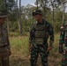 Three military personnel are standing together in an outdoor wooded area, appearing to be engaged in friendly conversation. One soldier is wearing US Army multicam uniform with a name tape reading 'VASQUEZ' and an American flag patch, while the other two are wearing Indonesian Army (TNI - Tentara Nasional Indonesia) camouflage uniforms with Indonesian flag patches. This appears to be a joint military exercise or cooperative engagement between US and Indonesian armed forces.