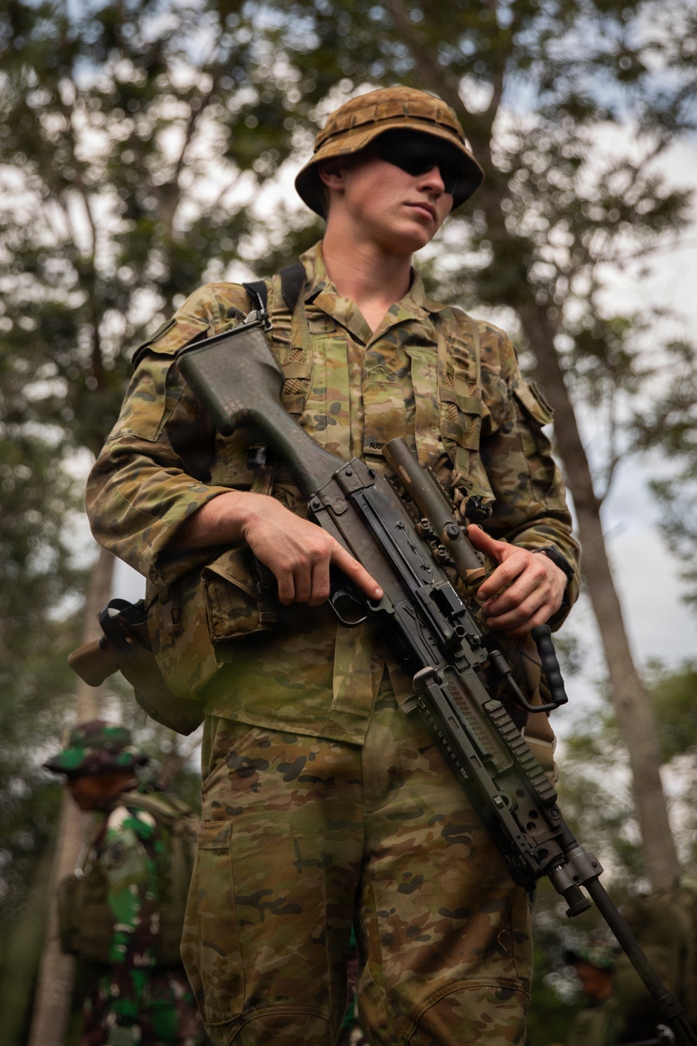 A soldier in Australian Defence Force (ADF) Multicam/AMCU camouflage uniform is standing in a wooded area holding a weapon, likely an EF88/F90 Austeyr assault rifle with an underslung grenade launcher. In the background, a second soldier wearing green Indonesian TNI (Tentara Nasional Indonesia) camouflage uniform is partially visible, suggesting a joint military exercise. The setting appears to be a forested training environment.