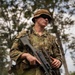 A soldier in Australian Defence Force (ADF) Multicam/AMCU camouflage uniform is standing in a wooded area holding a weapon, likely an EF88/F90 Austeyr assault rifle with an underslung grenade launcher. In the background, a second soldier wearing green Indonesian TNI (Tentara Nasional Indonesia) camouflage uniform is partially visible, suggesting a joint military exercise. The setting appears to be a forested training environment.