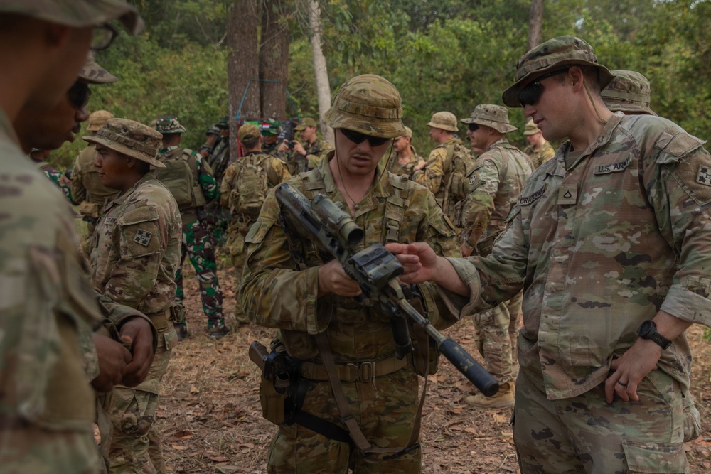 A group of military personnel in camouflage uniforms are gathered in a forested outdoor area, appearing to be engaged in a joint training exercise. Two soldiers in the foreground are examining a weapon, with one soldier holding what appears to be an M203 grenade launcher-equipped rifle. The presence of both U.S. Army personnel (identified by 'U.S. ARMY' tape) and soldiers in different camouflage patterns suggests a multinational military exercise.