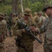 A group of military personnel in camouflage uniforms are gathered in a forested outdoor area, appearing to be engaged in a joint training exercise. Two soldiers in the foreground are examining a weapon, with one soldier holding what appears to be an M203 grenade launcher-equipped rifle. The presence of both U.S. Army personnel (identified by 'U.S. ARMY' tape) and soldiers in different camouflage patterns suggests a multinational military exercise.