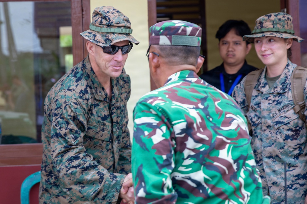 A military handshake meeting between a U.S. Marine in MARPAT digital camouflage and a soldier wearing green/brown camouflage consistent with Indonesian Armed Forces (TNI) uniform. A second U.S. Marine in MARPAT and a civilian with a blue lanyard badge observe in the background. The setting appears to be an outdoor or semi-outdoor facility, suggesting a bilateral military engagement or liaison visit.