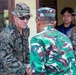 A military handshake meeting between a U.S. Marine in MARPAT digital camouflage and a soldier wearing green/brown camouflage consistent with Indonesian Armed Forces (TNI) uniform. A second U.S. Marine in MARPAT and a civilian with a blue lanyard badge observe in the background. The setting appears to be an outdoor or semi-outdoor facility, suggesting a bilateral military engagement or liaison visit.