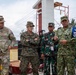A group of military personnel from multiple nations are gathered outdoors in what appears to be a joint military operation or humanitarian mission briefing. The group includes U.S. Army, U.S. Marine Corps, and Indonesian Armed Forces (TNI AU) personnel engaged in discussion. The setting appears to be a construction or disaster relief site based on visible building materials and infrastructure.