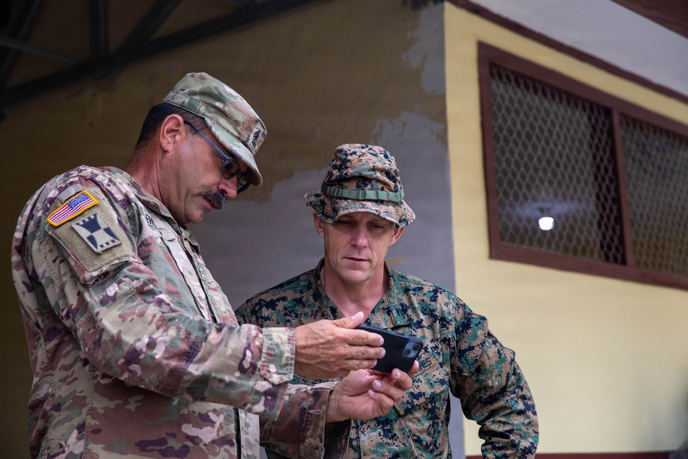 Two U.S. military personnel in different camouflage uniforms are examining a smartphone together outdoors near a building. The individual on the left wears U.S. Army OCP (Operational Camouflage Pattern) uniform with a U.S. flag patch and rank insignia, while the individual on the right wears U.S. Marine Corps MARPAT digital camouflage with a boonie hat. They appear to be reviewing information or imagery on the device.