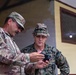 Two U.S. military personnel in different camouflage uniforms are examining a smartphone together outdoors near a building. The individual on the left wears U.S. Army OCP (Operational Camouflage Pattern) uniform with a U.S. flag patch and rank insignia, while the individual on the right wears U.S. Marine Corps MARPAT digital camouflage with a boonie hat. They appear to be reviewing information or imagery on the device.