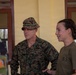 Two individuals in a military context are engaged in conversation inside a building with yellow walls and wooden-framed windows. The person on the left is wearing U.S. Marines MARPAT woodland camouflage uniform with a boonie cover, while the person on the right is wearing an olive drab military t-shirt. A third individual in a different camouflage pattern is partially visible on the left edge of the frame.