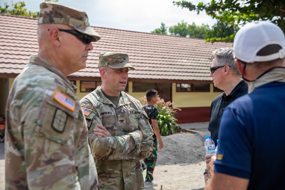 Two U.S. Army soldiers in MultiCam/OCP camouflage uniforms are engaged in conversation with two civilians outdoors near a building with a red tile roof. One soldier's name tape reads 'LAWRENCE' and both wear U.S. Army insignia with American flag patches. The setting appears to be an overseas location, possibly Southeast Asia, based on architectural style and a partially visible individual in the background wearing what appears to be Indonesian military uniform.
