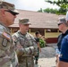 Two U.S. Army soldiers in MultiCam/OCP camouflage uniforms are engaged in conversation with two civilians outdoors near a building with a red tile roof. One soldier's name tape reads 'LAWRENCE' and both wear U.S. Army insignia with American flag patches. The setting appears to be an overseas location, possibly Southeast Asia, based on architectural style and a partially visible individual in the background wearing what appears to be Indonesian military uniform.