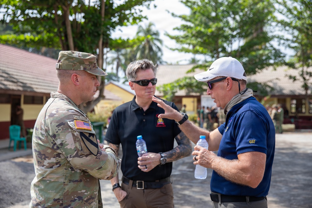 Three men are engaged in a conversation outdoors at what appears to be a military or government compound in a tropical setting. One man is wearing U.S. Army combat uniform with an American flag patch, while the other two are in civilian attire. The setting and context suggest a military liaison or advisory meeting.