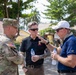 Three men are engaged in a conversation outdoors at what appears to be a military or government compound in a tropical setting. One man is wearing U.S. Army combat uniform with an American flag patch, while the other two are in civilian attire. The setting and context suggest a military liaison or advisory meeting.