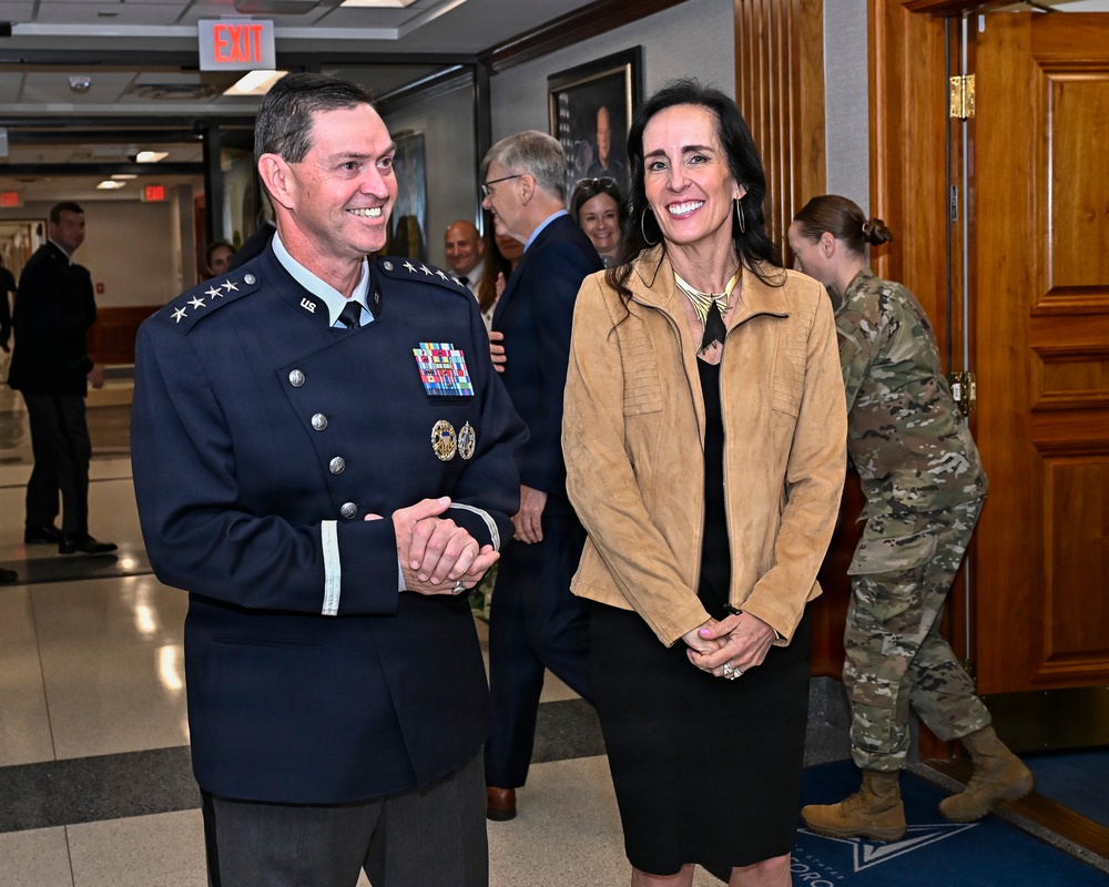 A high-ranking U.S. Air Force general in dress uniform stands alongside a civilian woman in a hallway of what appears to be a military facility. Both are smiling and appear to be at an official event or ceremony. Several other individuals, including a uniformed service member in camouflage, are visible in the background.