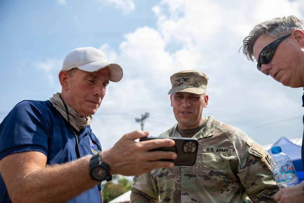 Three men are gathered outdoors reviewing content on a smartphone. One man in U.S. Army camouflage uniform with a colonel's eagle insignia is flanked by two civilians, one wearing a white cap and blue polo shirt and another wearing sunglasses. The setting appears to be an outdoor operational or relief site with a blue tent visible in the background.