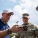 Three men are gathered outdoors reviewing content on a smartphone. One man in U.S. Army camouflage uniform with a colonel's eagle insignia is flanked by two civilians, one wearing a white cap and blue polo shirt and another wearing sunglasses. The setting appears to be an outdoor operational or relief site with a blue tent visible in the background.