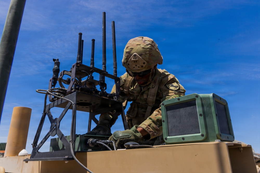 A soldier in full combat gear is working on top of a military vehicle, appearing to configure or inspect electronic equipment. The soldier is wearing OCP (Operational Camouflage Pattern) uniform, a tactical helmet with face protection, and gloves. Multiple antennas and what appears to be an electronic warfare or communications system are mounted on the vehicle.