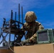 A soldier in full combat gear is working on top of a military vehicle, appearing to configure or inspect electronic equipment. The soldier is wearing OCP (Operational Camouflage Pattern) uniform, a tactical helmet with face protection, and gloves. Multiple antennas and what appears to be an electronic warfare or communications system are mounted on the vehicle.