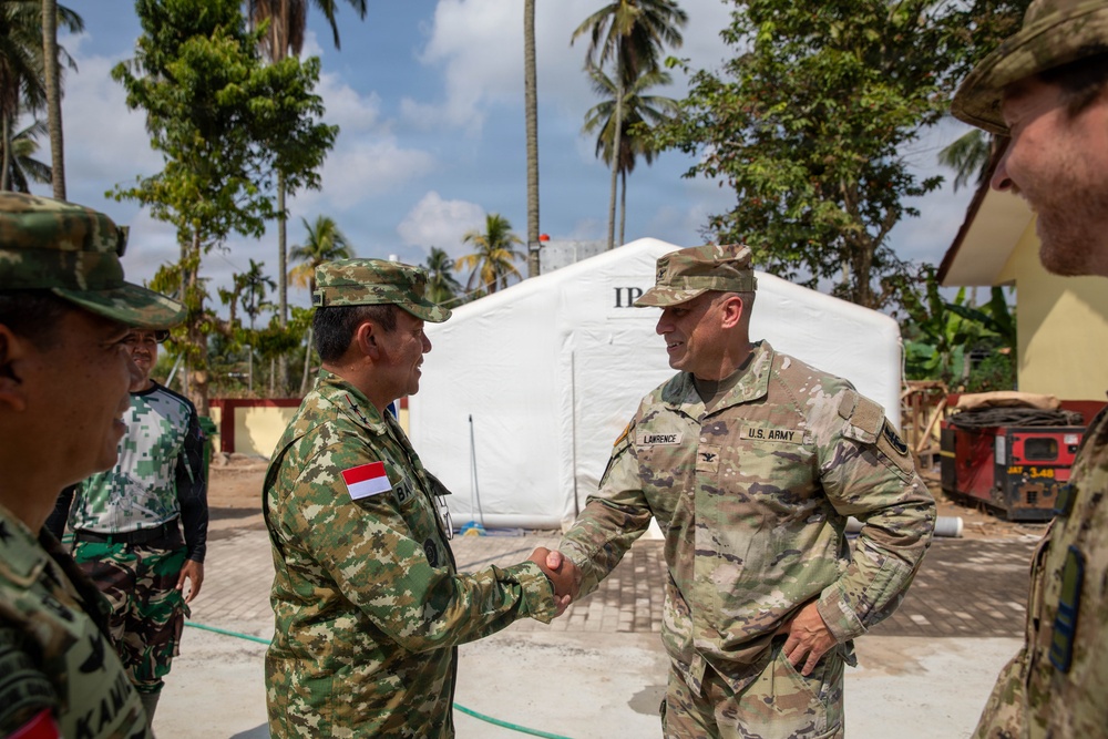 A U.S. Army soldier named Lawrence is shaking hands with an Indonesian military officer in what appears to be a field or disaster relief setting. The meeting takes place outdoors near a white tent structure, likely a temporary command or medical facility, surrounded by palm trees suggesting a tropical location. Additional military personnel from both forces are visible in the background.