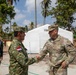A U.S. Army soldier named Lawrence is shaking hands with an Indonesian military officer in what appears to be a field or disaster relief setting. The meeting takes place outdoors near a white tent structure, likely a temporary command or medical facility, surrounded by palm trees suggesting a tropical location. Additional military personnel from both forces are visible in the background.