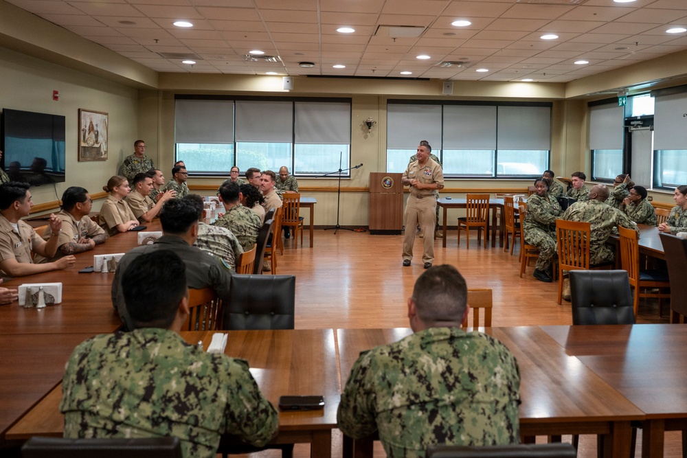 A military briefing or meeting is taking place in a formal conference room, with a senior officer in tan/khaki service uniform standing at a podium addressing a group of approximately 30 military personnel. The audience is seated at wooden tables arranged in a U-shape or classroom configuration, wearing a mix of Navy Working Uniform (NWU) camouflage and khaki service uniforms. The setting appears to be a military installation conference or meeting room.