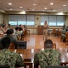 A military briefing or meeting is taking place in a formal conference room, with a senior officer in tan/khaki service uniform standing at a podium addressing a group of approximately 30 military personnel. The audience is seated at wooden tables arranged in a U-shape or classroom configuration, wearing a mix of Navy Working Uniform (NWU) camouflage and khaki service uniforms. The setting appears to be a military installation conference or meeting room.