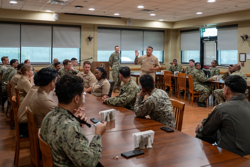 A military briefing or meeting is taking place in what appears to be a dining facility or conference room on a military installation. Two senior officers in tan/khaki uniforms are standing and addressing a group of approximately 25-30 Navy personnel seated around tables arranged in a U-shape. The attendees are wearing a mix of Navy Working Uniform (NWU) digital camouflage and khaki service uniforms.