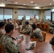 A military briefing or meeting is taking place in what appears to be a dining facility or conference room on a military installation. Two senior officers in tan/khaki uniforms are standing and addressing a group of approximately 25-30 Navy personnel seated around tables arranged in a U-shape. The attendees are wearing a mix of Navy Working Uniform (NWU) digital camouflage and khaki service uniforms.