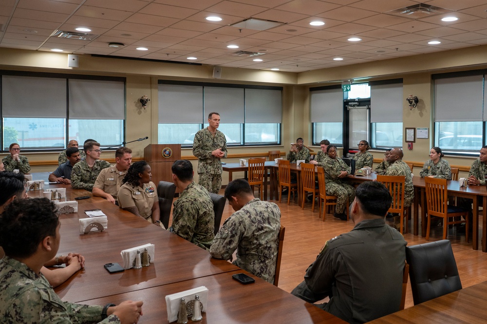 A military briefing or meeting is taking place in a formal conference/dining room, with one standing officer addressing a group of seated personnel in military uniforms. The majority of attendees appear to be U.S. Navy personnel in NWU (Navy Working Uniform) camouflage, with at least one individual in khaki service uniform. The setting appears to be a military installation facility, possibly a wardroom or conference room.