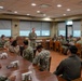 A military briefing or meeting is taking place in a formal conference/dining room, with one standing officer addressing a group of seated personnel in military uniforms. The majority of attendees appear to be U.S. Navy personnel in NWU (Navy Working Uniform) camouflage, with at least one individual in khaki service uniform. The setting appears to be a military installation facility, possibly a wardroom or conference room.
