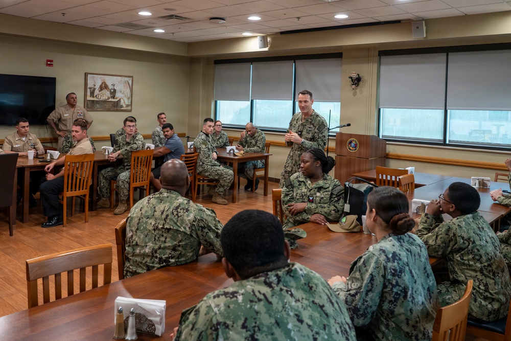 A military briefing or meeting is taking place in a formal conference/dining room setting. A standing officer in U.S. Navy digital camouflage (NWU Type III) uniform appears to be addressing a group of seated sailors. The attendees are arranged at wooden tables in what appears to be a Navy installation meeting room.