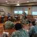 A military briefing or meeting is taking place in a formal conference/dining room setting. A standing officer in U.S. Navy digital camouflage (NWU Type III) uniform appears to be addressing a group of seated sailors. The attendees are arranged at wooden tables in what appears to be a Navy installation meeting room.