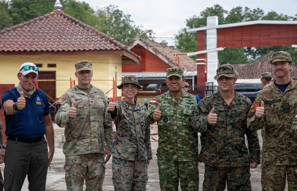 A group of six military personnel and one civilian pose together giving thumbs-up gestures in what appears to be a multinational military exercise or joint operation. The group includes members from at least two identified forces: the U.S. Army (name tape 'Lawrence' visible), U.S. Marines, and Indonesian military (TNI AU - Indonesian Air Force, with name tape 'Bambang' visible). They are standing in front of buildings with terracotta-tiled roofs consistent with Southeast Asian architecture.
