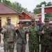 A group of six military personnel and one civilian pose together giving thumbs-up gestures in what appears to be a multinational military exercise or joint operation. The group includes members from at least two identified forces: the U.S. Army (name tape 'Lawrence' visible), U.S. Marines, and Indonesian military (TNI AU - Indonesian Air Force, with name tape 'Bambang' visible). They are standing in front of buildings with terracotta-tiled roofs consistent with Southeast Asian architecture.