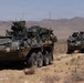 The image shows a column of at least two wheeled armored fighting vehicles traversing arid desert terrain with mountains visible in the background. The vehicles appear to be LAV-type or Stryker-type 8-wheeled armored personnel carriers equipped with weapon systems and communications equipment. Personnel are visible in the turret hatches of the vehicles, suggesting an active patrol or convoy operation.