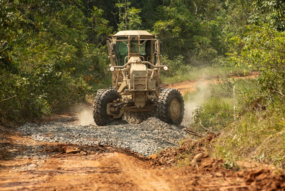A military-colored (tan/beige) motor grader is actively grading and compacting gravel onto a dirt road carved through a dense tropical or subtropical forest. The machine appears to be constructing or improving a road surface by spreading crushed stone aggregate over the red clay soil. Dust is visible around the rear wheels, indicating active operation.