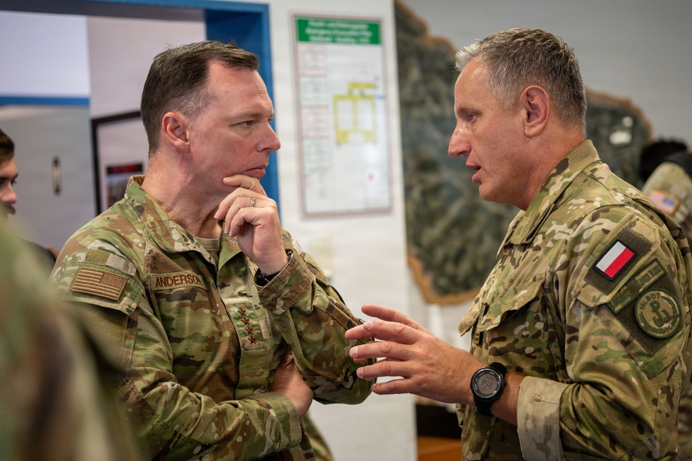 Two military officers in MultiCam/OCP camouflage uniforms are engaged in a focused conversation. The officer on the left, identified by a name tape reading 'ANDERSON' and wearing four stars indicating a General rank, is listening attentively with hand raised to chin. The officer on the right wears a Polish flag patch and a unit patch, suggesting a multinational military meeting or briefing.